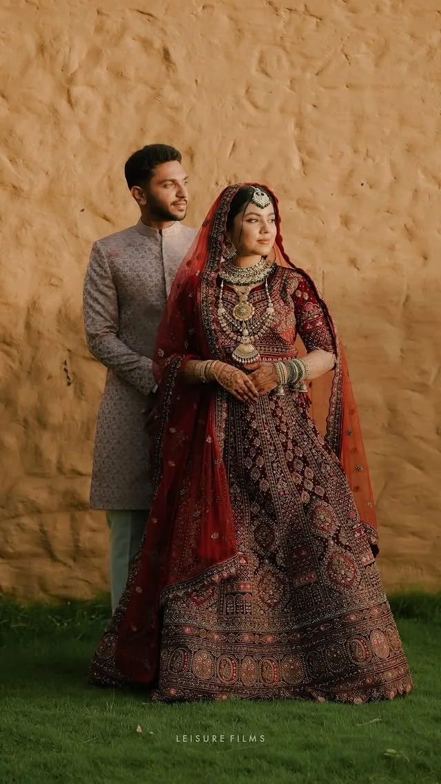 Elegant bride in a heavily embroidered red lehenga with statement jewelry and groom in a sherwani, standing against a rustic wall, beautifully captured by the best wedding photographer in Calicut
