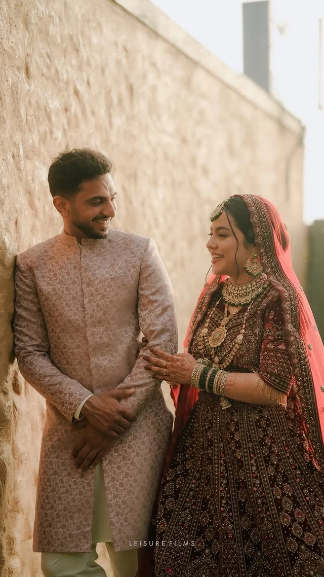 A bride in a heavily embellished red lehenga smiles lovingly at her groom in a pastel sherwani as they share a candid moment against a rustic wal beautifully captured by the best wedding photographer in Calicut.