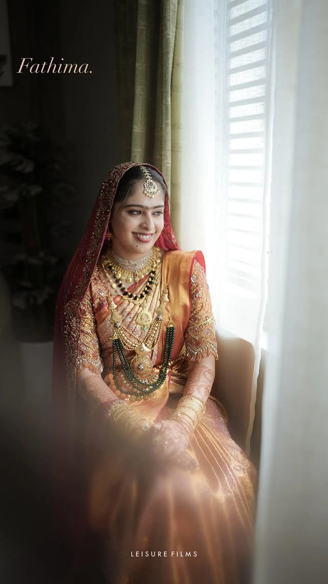 Beautiful bride in a golden silk saree with red dupatta, adorned with layered gold and green jewelry, smiling by the window, captured by the best wedding photographer in Calicut.