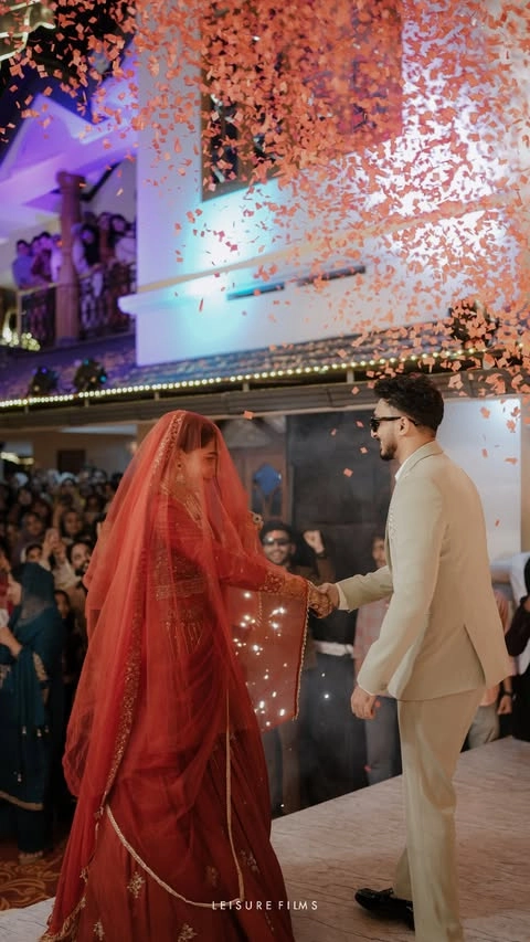 A bride in a stunning red gown with a veil holds her groom’s hand under a shower of flower petals, surrounded by cheering guests a magical moment beautifully captured by the best wedding photographer in Calicut.